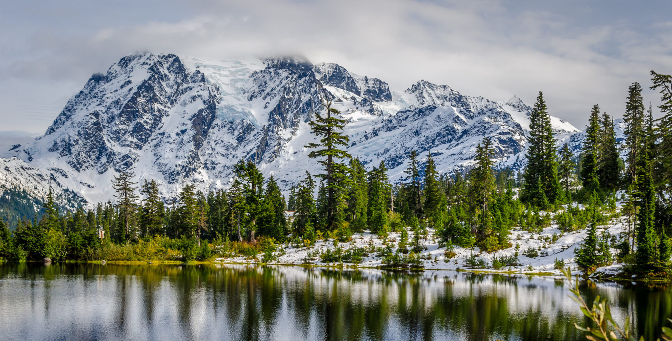 A lake reflects the image of a snow covered mountain and moraine lined with evergreen trees.